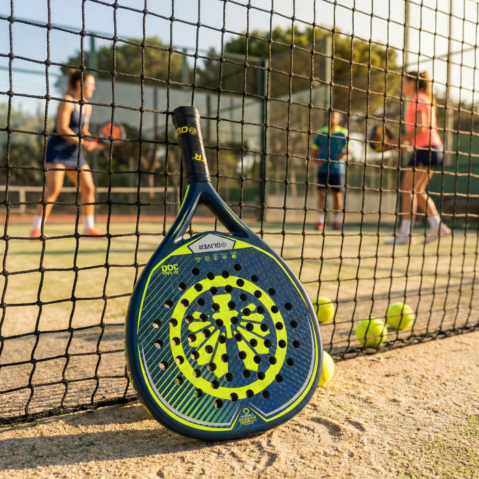 Der OLIVER Padel-Schläger Air Max 300, blau mit gelbem Design, lehnt an einem Padel Netz, im Hintergrund spielen Personen Padel.